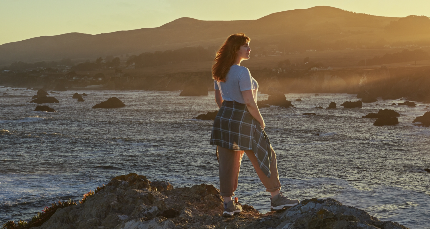 Woman standing by rocky shore at sunset mobile