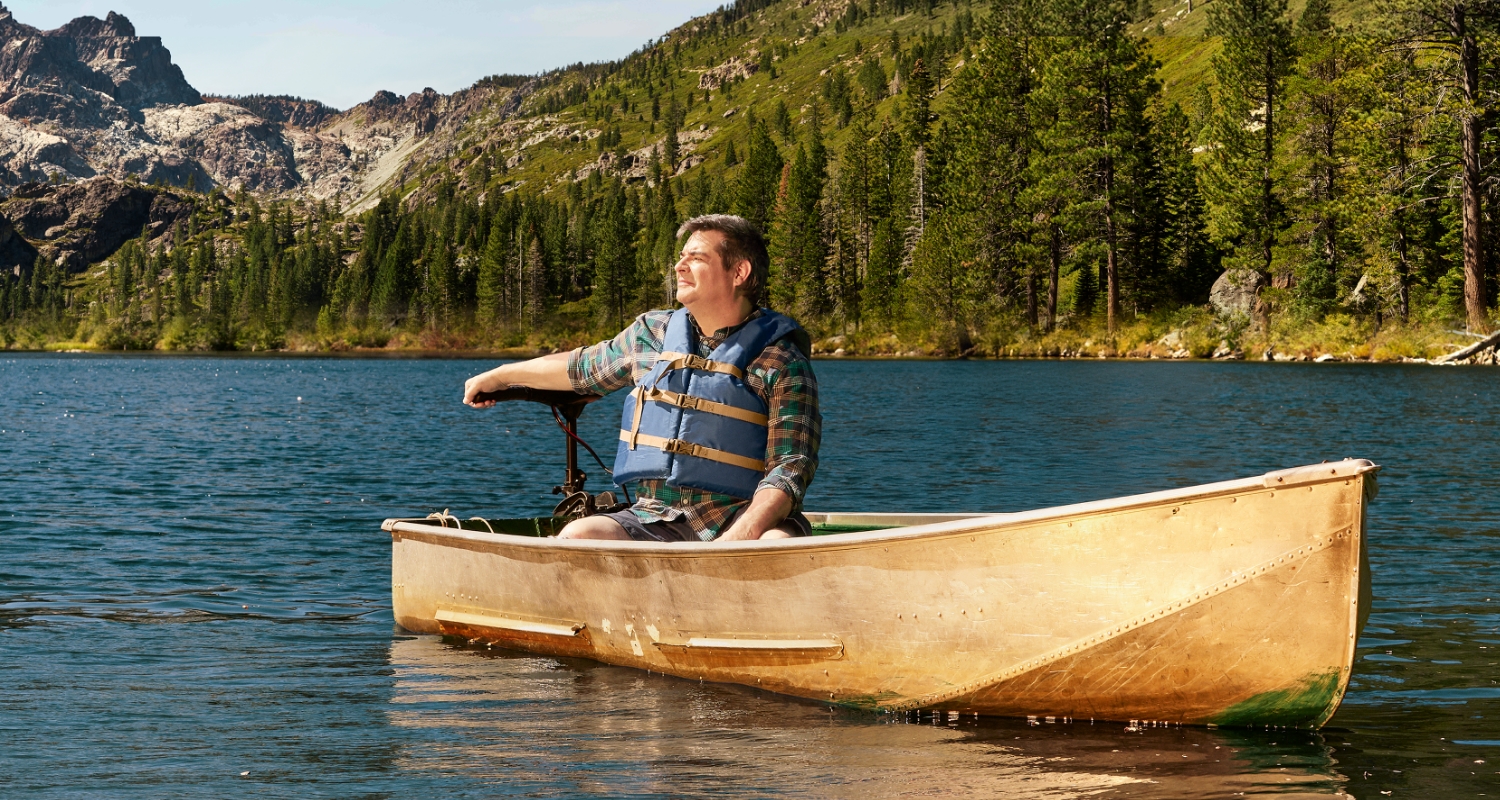 Man wearing a life vest sitting on a boat on a lake surrounded by nature