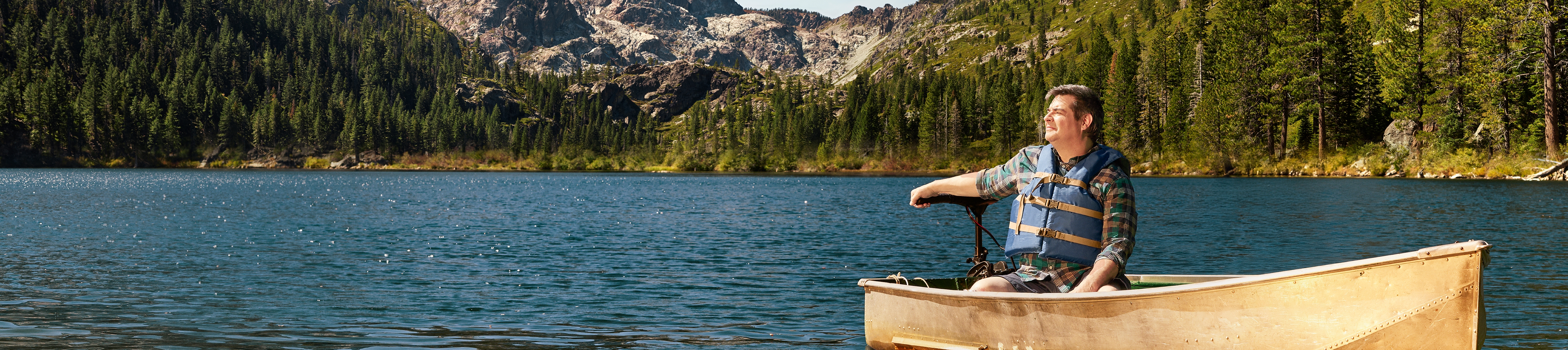 Man wearing a life vest sitting on a boat on a lake surrounded by nature