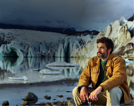 Man wearing a brown jacket and green sweater sitting by a lake with glaciers in the background