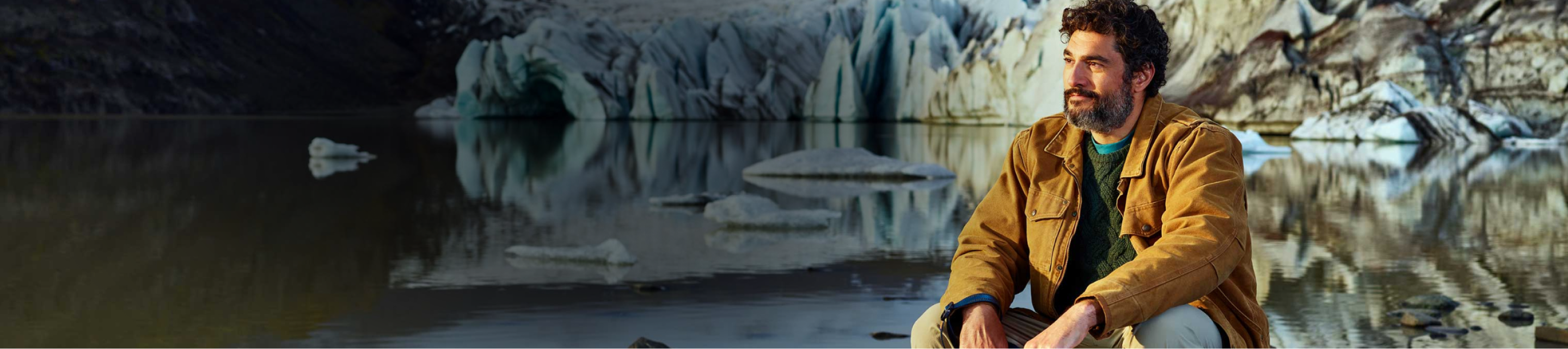 Man wearing a brown jacket and green sweater sitting by a lake with glaciers in the background