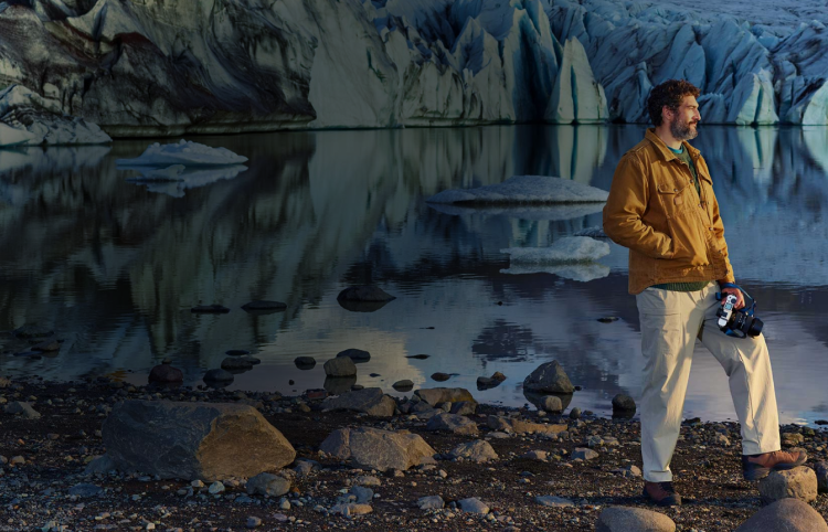 Man wearing a brown jacket holding a camera staring into the distance with a lake and glaciers behind him
