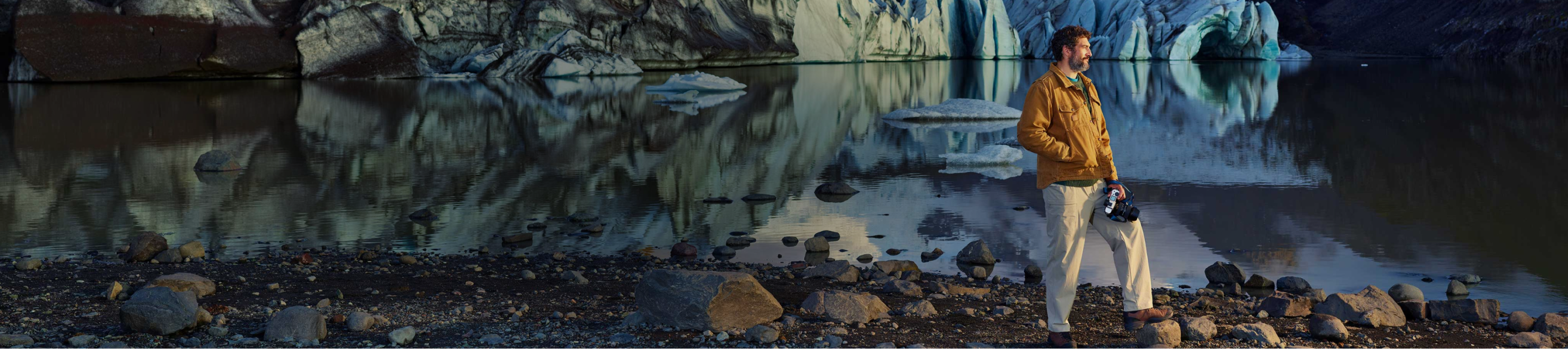Man wearing a brown jacket holding a camera staring into the distance with a lake and glaciers behind him
