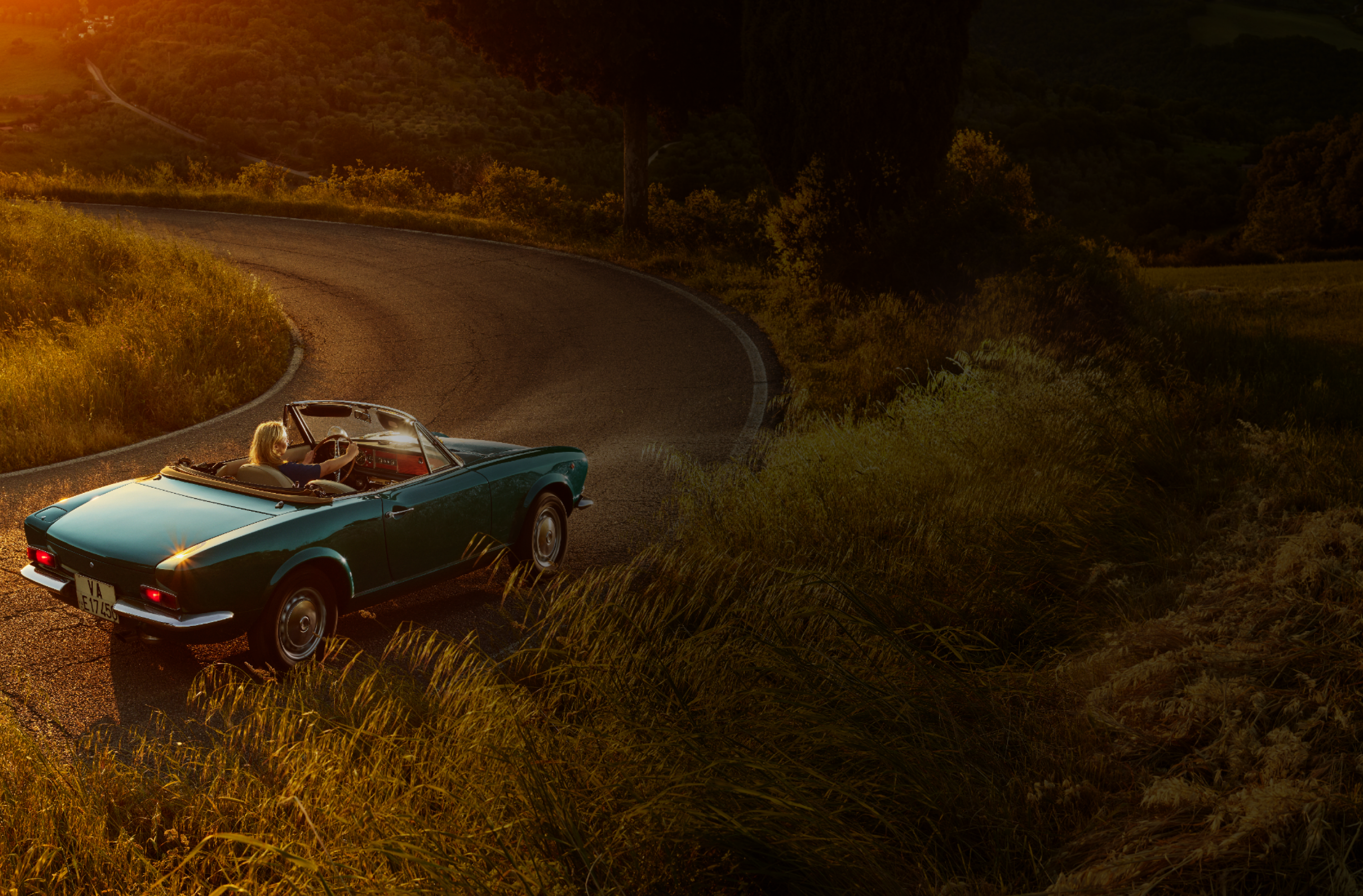 Blue convertible car with a woman driving on a winding road during sunset