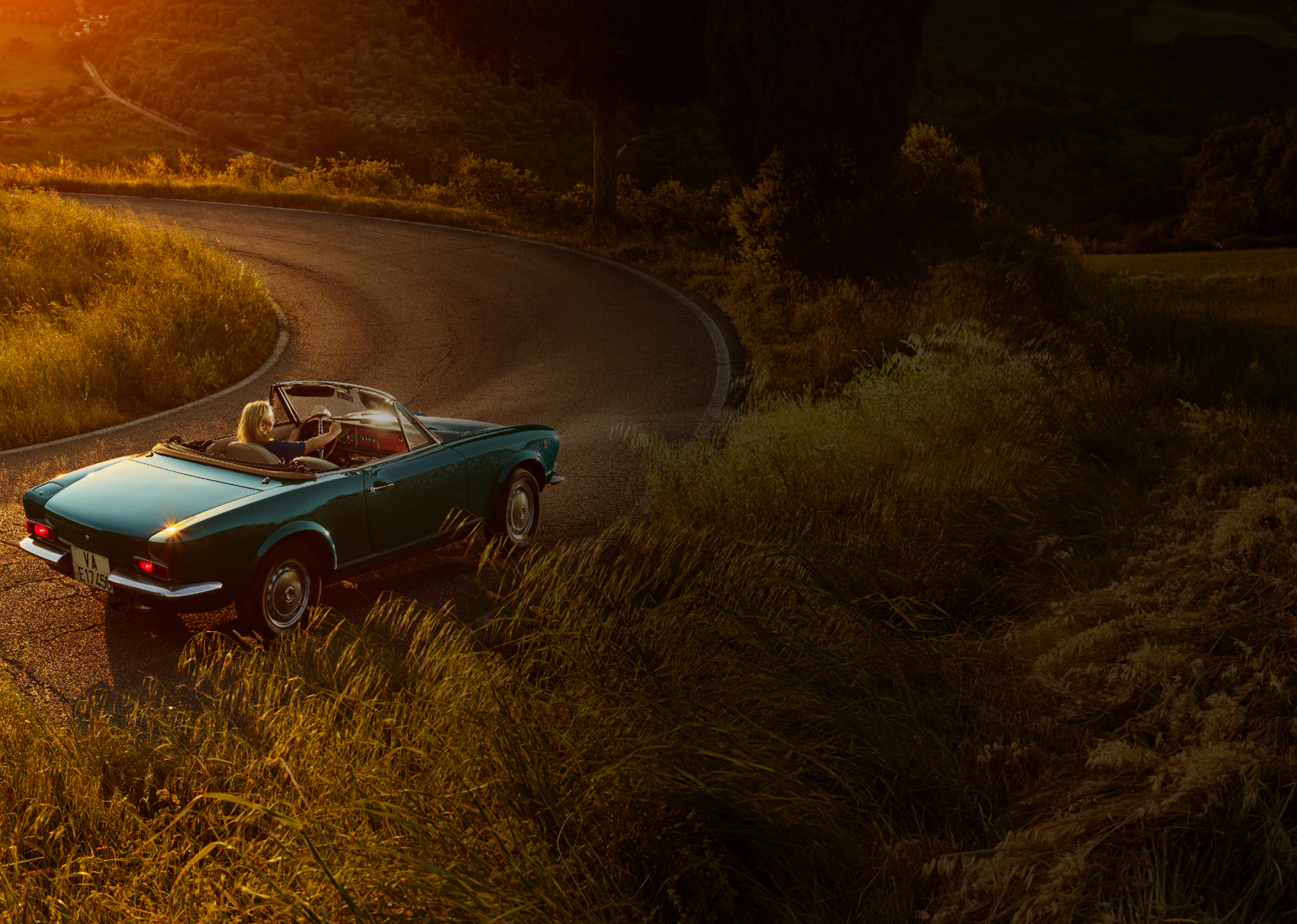 Blue convertible car with a woman driving on a winding road during sunset